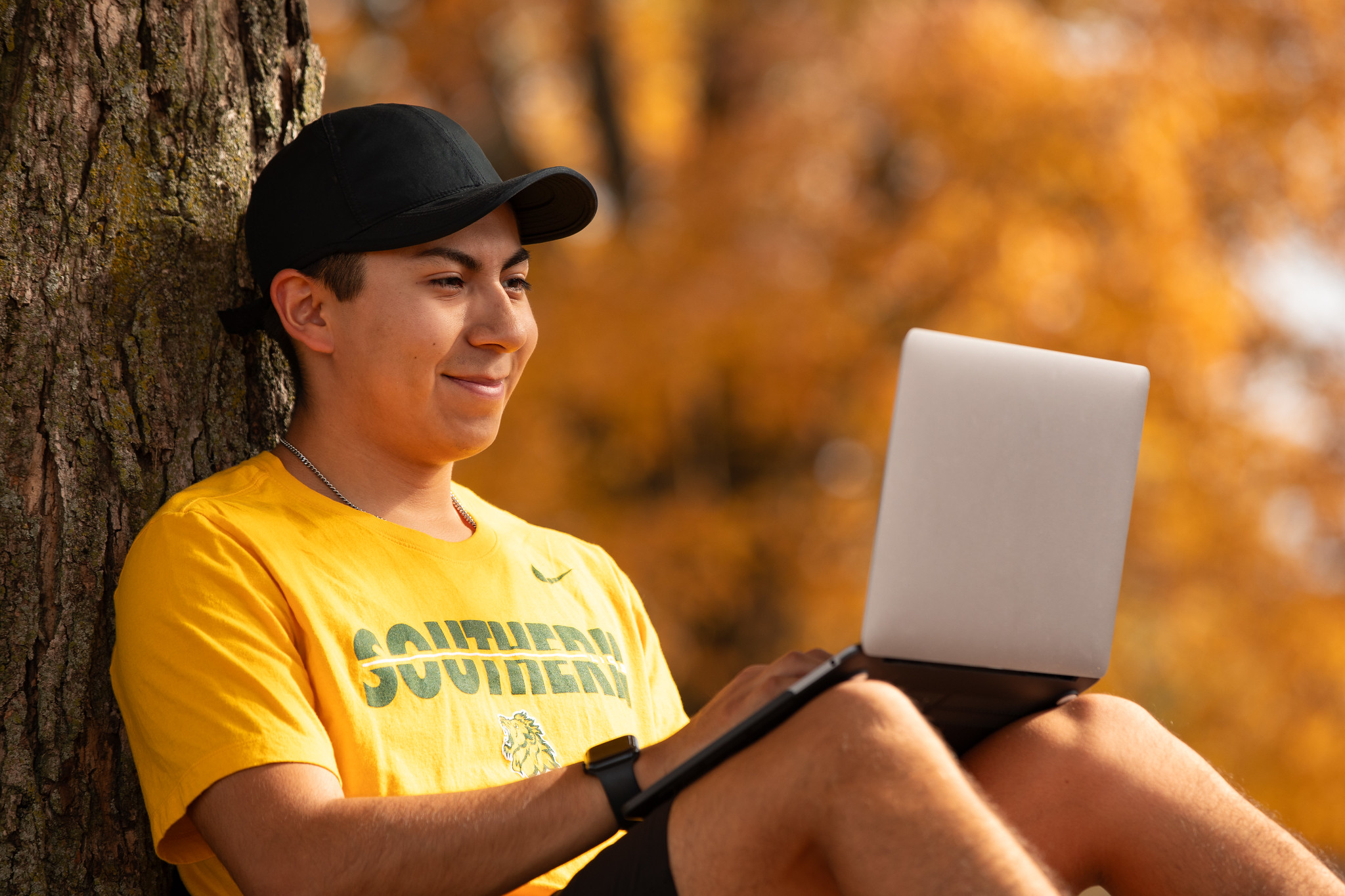A person sits outdoors leaning against a tree trunk, using a laptop placed on their lap. The person wears a yellow Missouri Southern T-shirt and a black cap. The setting includes autumn foliage with orange and yellow leaves, grass, and warm sunlight filtering through the trees. 