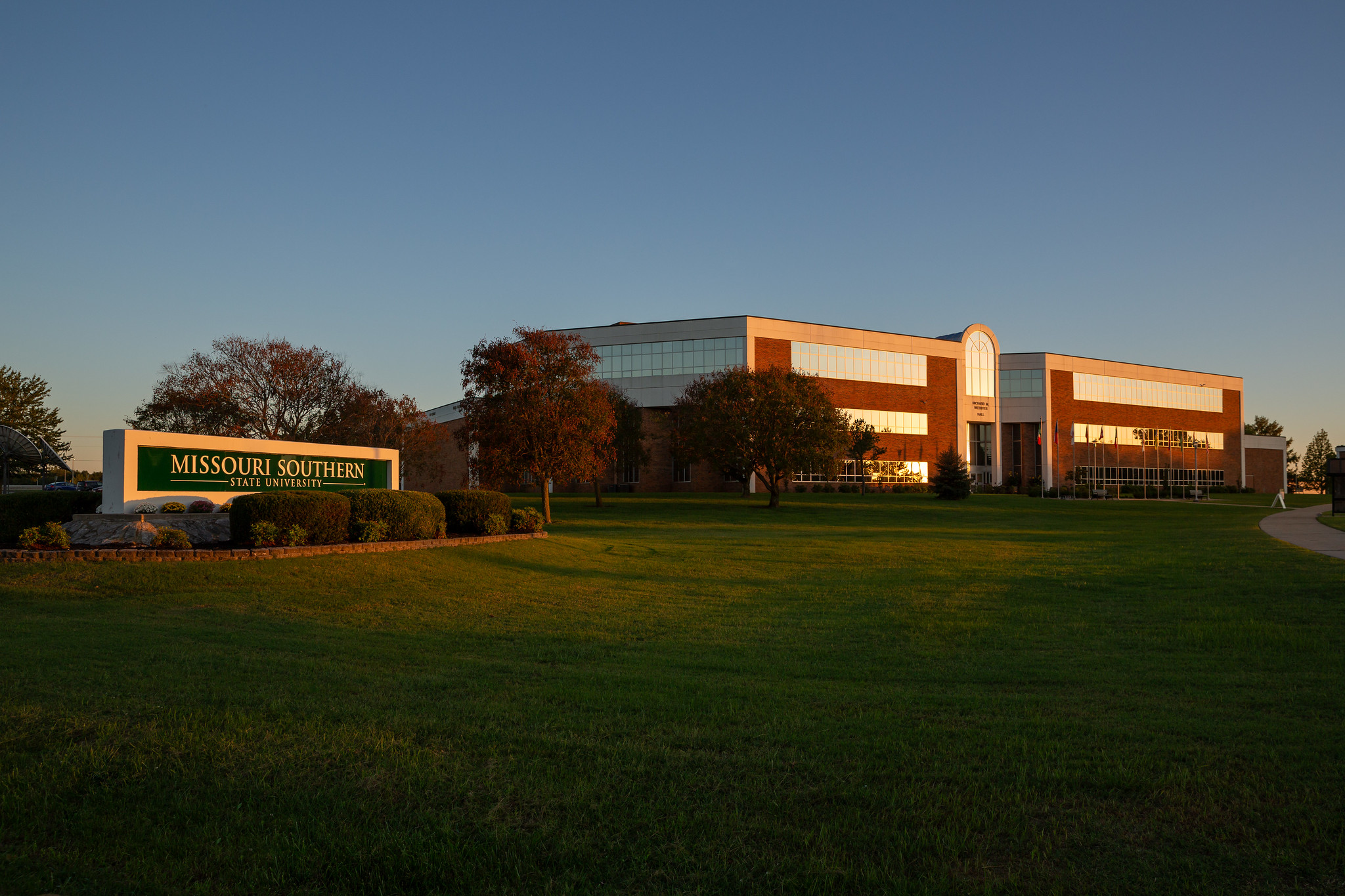 A wide outdoor view of the Missouri Southern State University campus at sunset. In the foreground, a manicured green lawn stretches toward a brick-and-glass academic building with large windows and a curved central entrance. To the left, a stone campus sign reads 'Missouri Southern State University.' Trees with autumn foliage line the grounds under warm evening light and a clear sky.