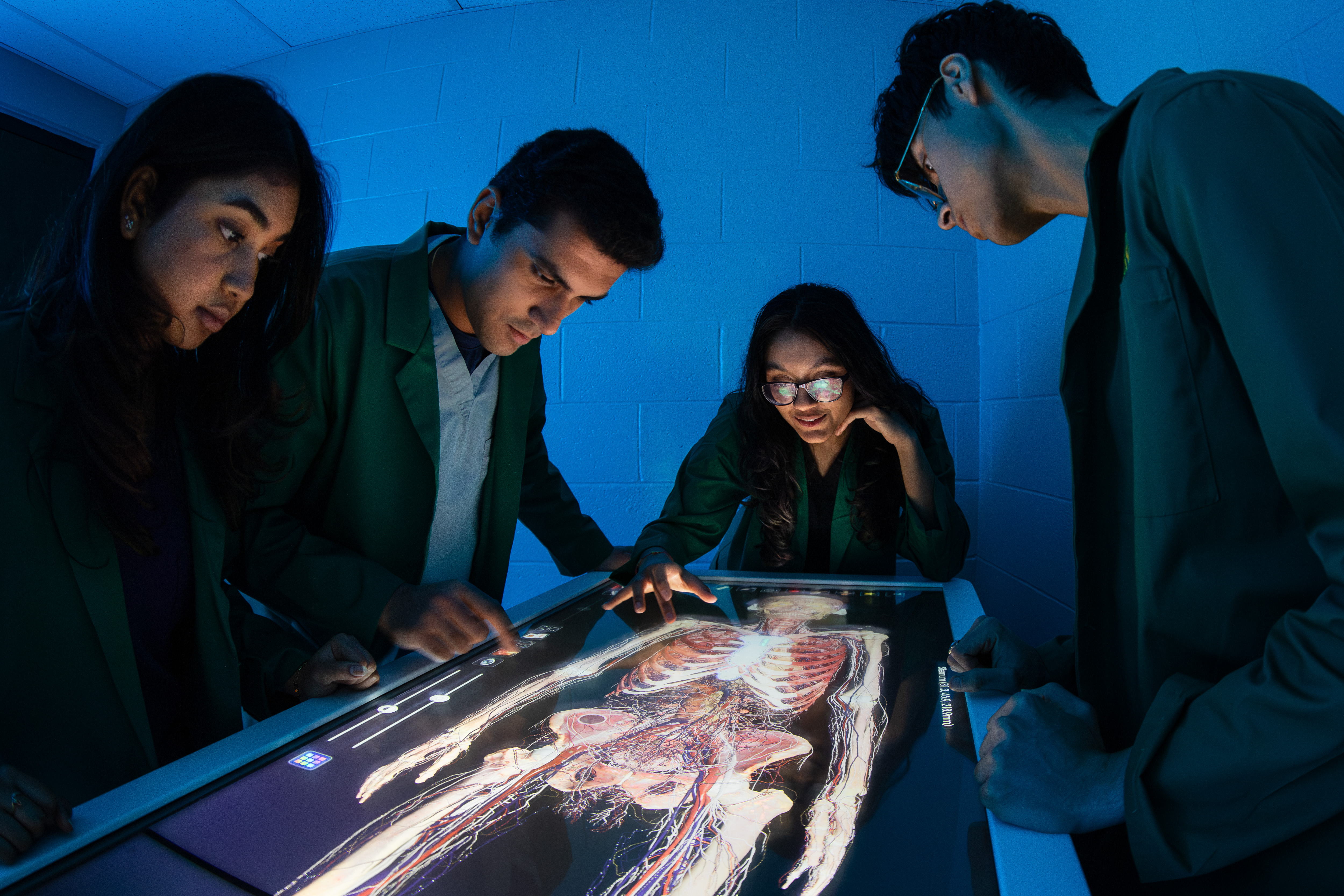 A group of people wearing lab coats gather around an illuminated digital anatomy table, interacting with a detailed 3D anatomical model displayed on the screen in a dimly lit room.