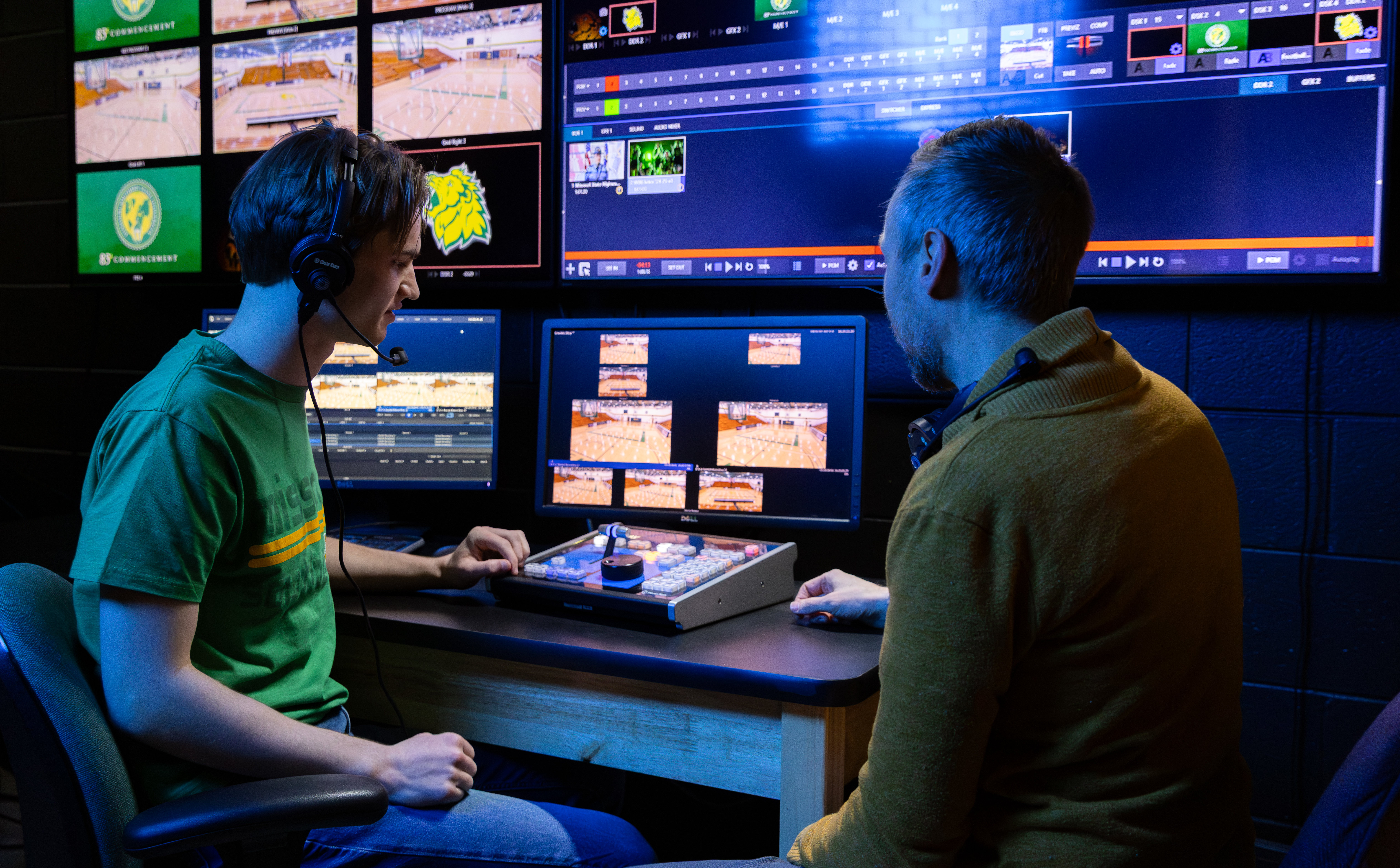 Two people sit at a video production control desk, wearing headsets and monitoring multiple screens displaying live sports footage while operating switching and editing equipment.