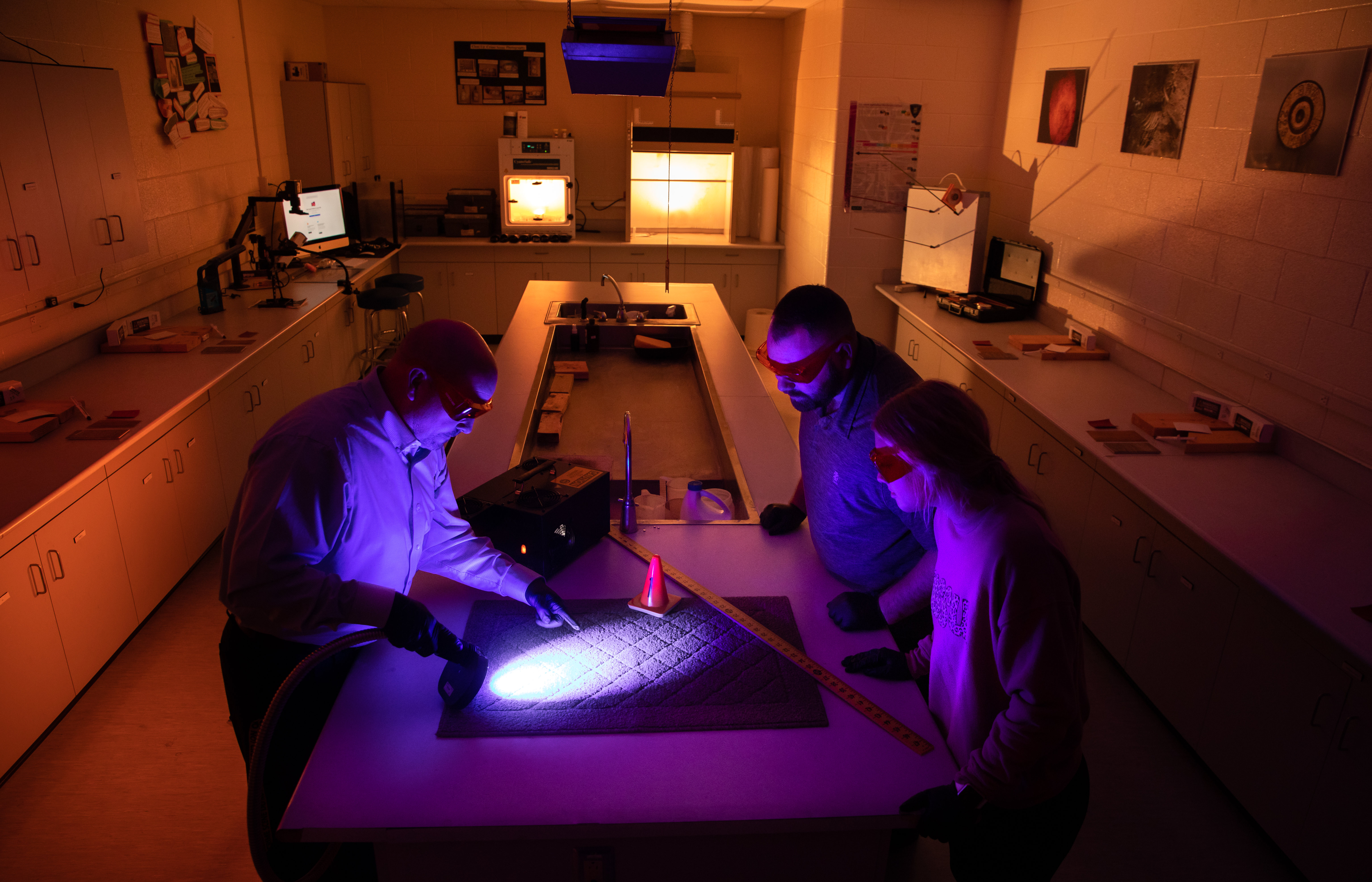 Three people stand around a laboratory table illuminated by purple and amber lighting, examining a large forensic-style surface with measuring tools and a spotlight while surrounded by lab equipment and computers.