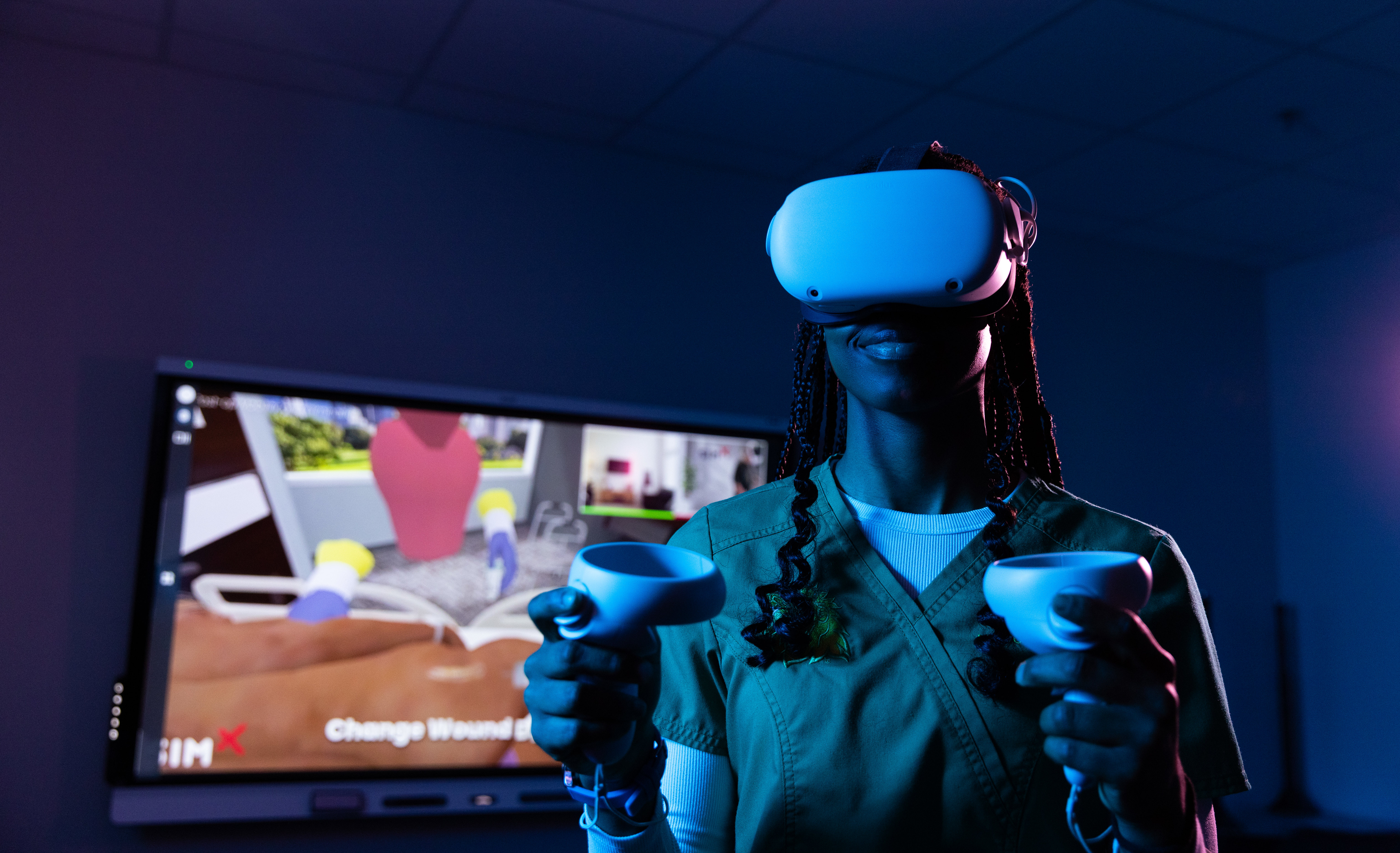 alt=A person wearing medical scrubs uses a virtual reality headset and two handheld controllers in a dimly lit room, with a large screen behind them showing a medical simulation interface.