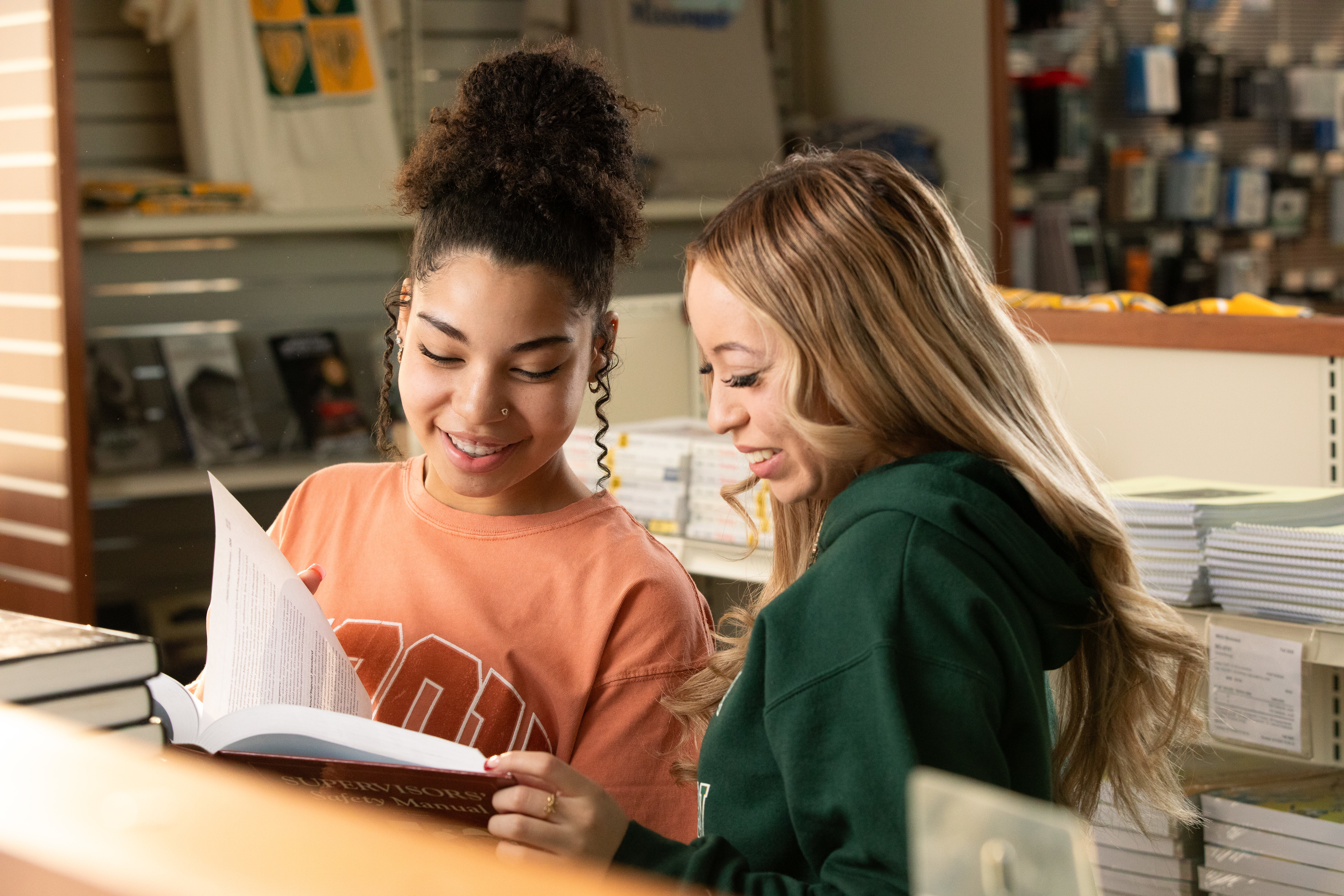 Two people standing in a campus bookstore look together at an open book, surrounded by shelves of textbooks and school merchandise.
