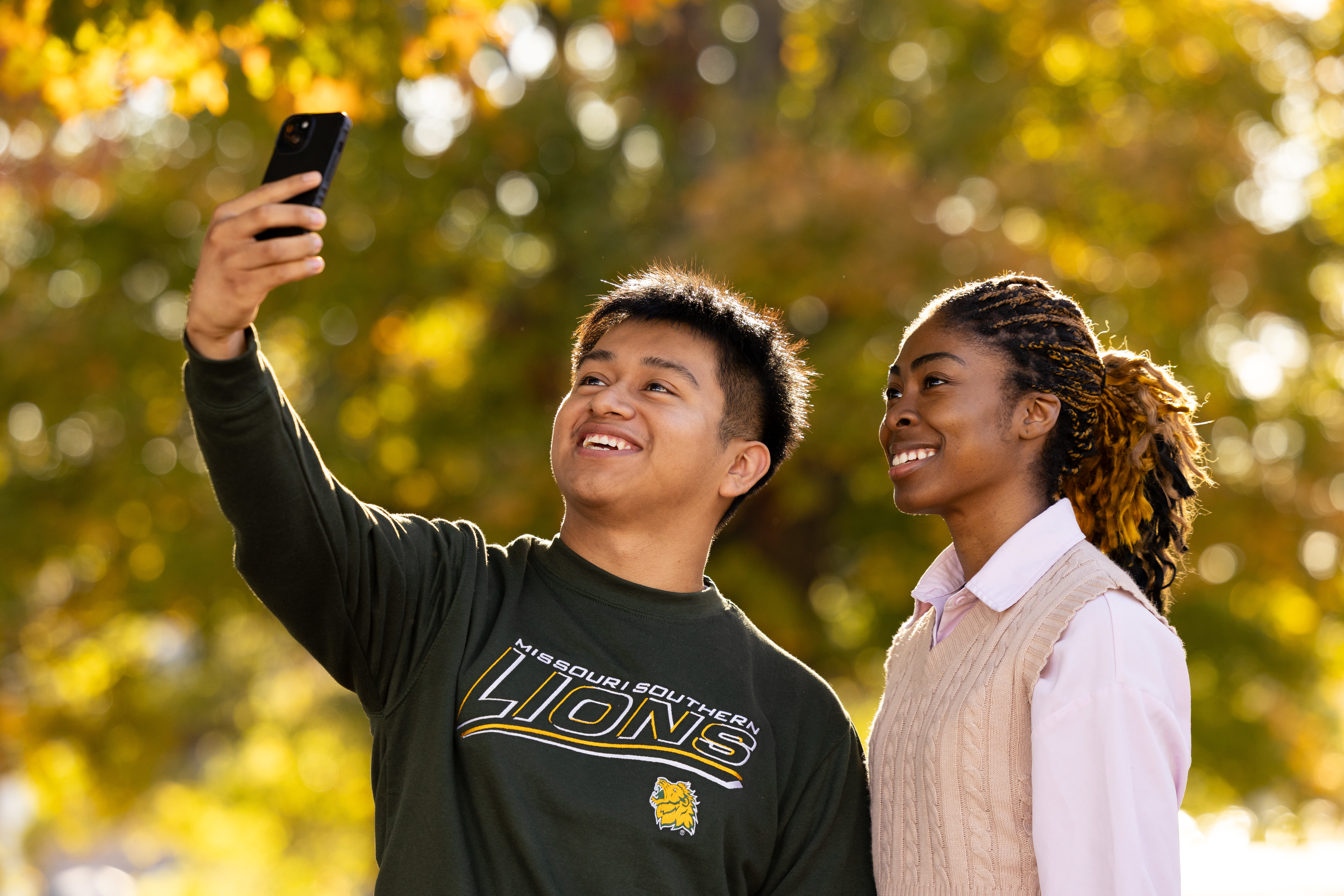 Two people standing outdoors in front of autumn foliage, with one holding up a smartphone to take a selfie while wearing a Missouri Southern Lions shirt.