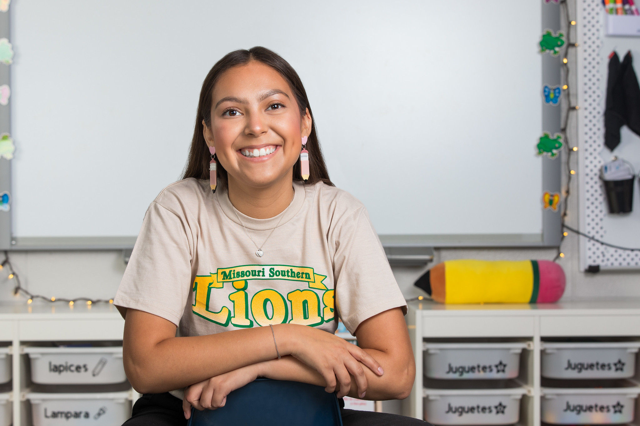 A person wearing a Missouri Southern Lions T‑shirt sits with arms crossed in a brightly lit classroom, with organized storage bins and a large pencil‑shaped decoration visible in the background.